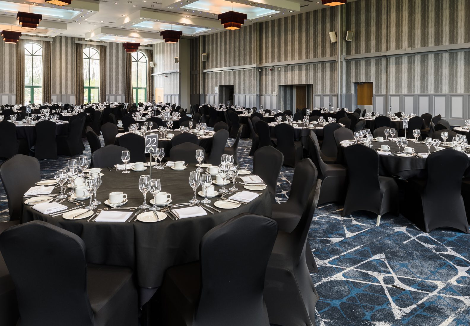 A banquet hall with round tables set with black tablecloths, glassware, and white napkins. Black chair covers and patterned carpet complete the room.