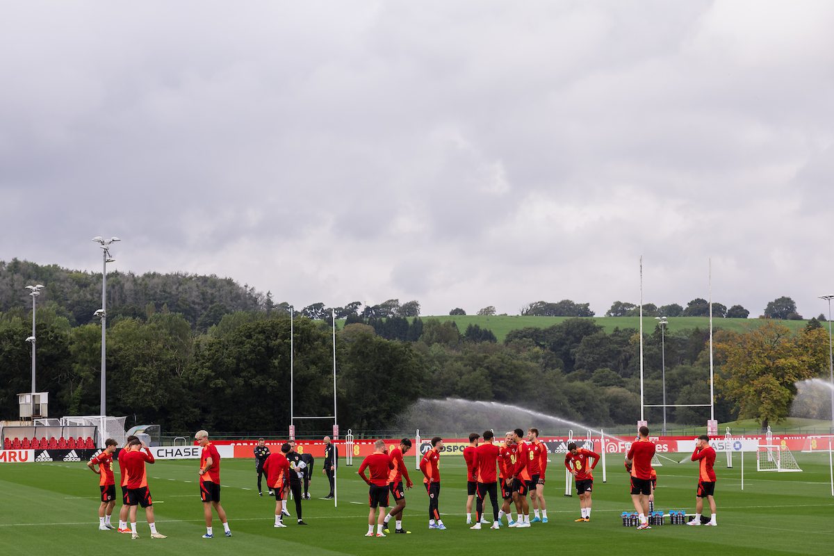 Soccer team in red jerseys gathered on a field, with sprinklers active in the background, surrounded by trees and a cloudy sky.
