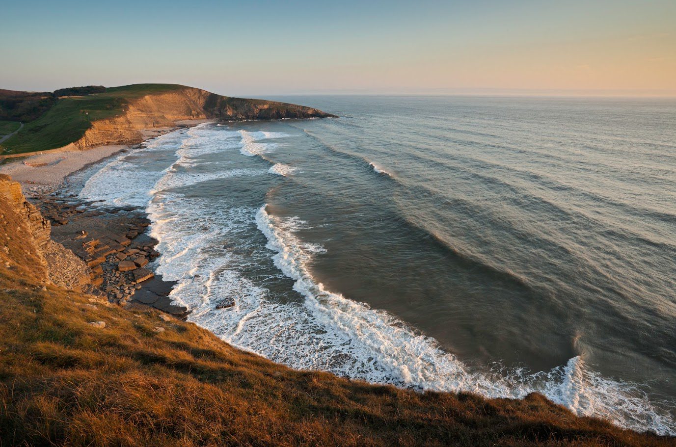 Waves crashing on a rocky coastline at sunset, with grassy cliffs in the foreground and a distant horizon.