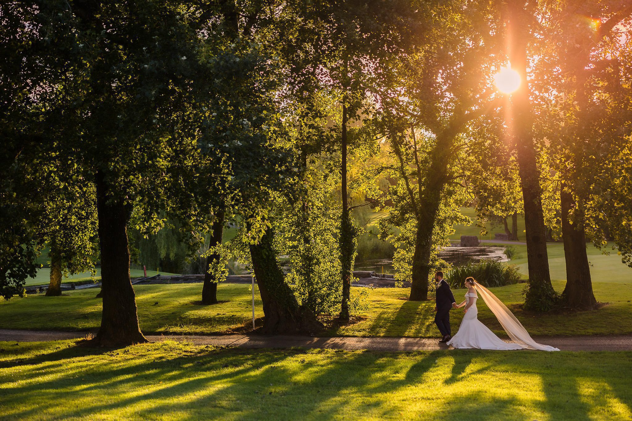 A bride and groom walk along a tree-lined path bathed in warm sunlight, with long shadows cast on the grass.