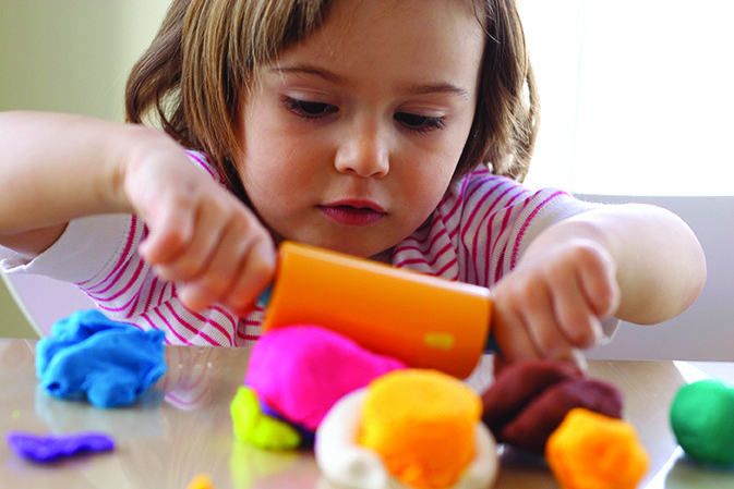 A child intently moulds colourful clay with a plastic tool on a table.