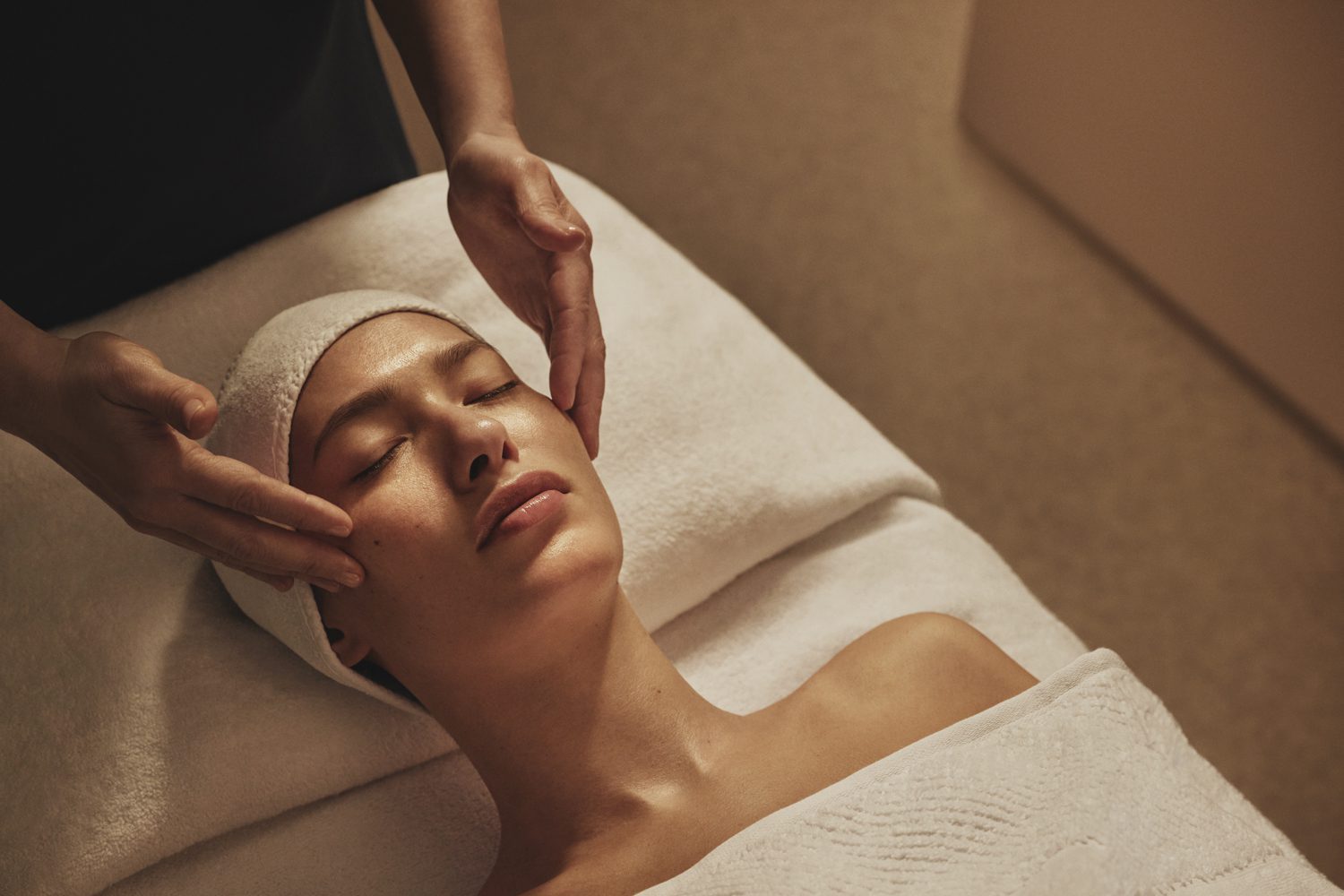 Person receiving a facial massage while lying on a spa table, eyes closed, covered with a towel, and head wrapped in a cloth.