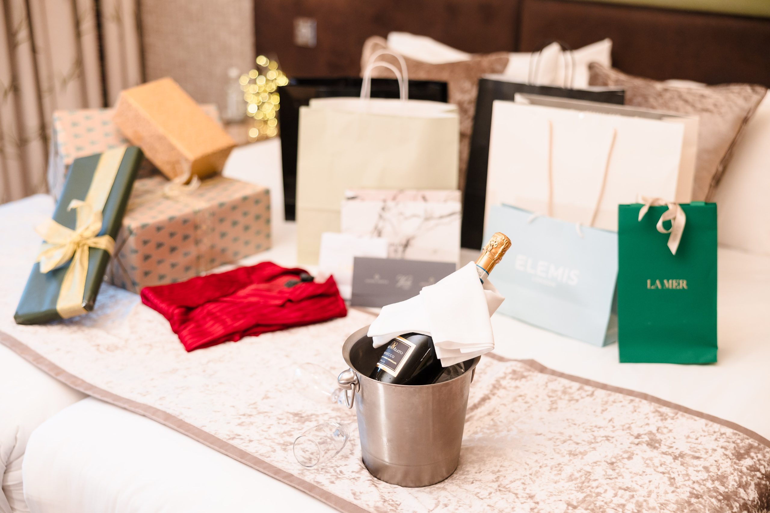 Gift bags, wrapped presents, and a bottle of wine in an ice bucket arranged on a bed in a hotel room.
