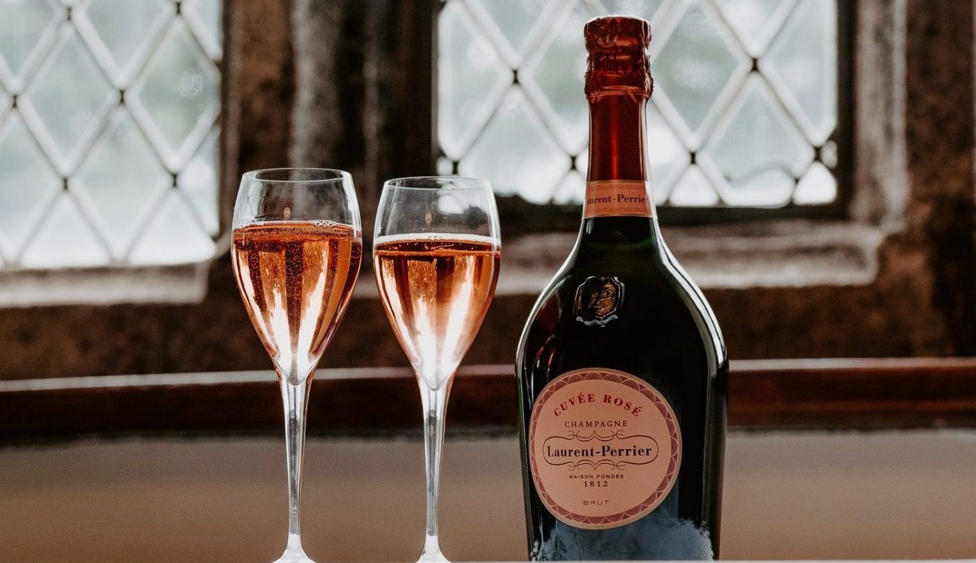 A bottle of Laurent-Perrier rosé champagne and two filled glasses are set on a white surface in front of a window.