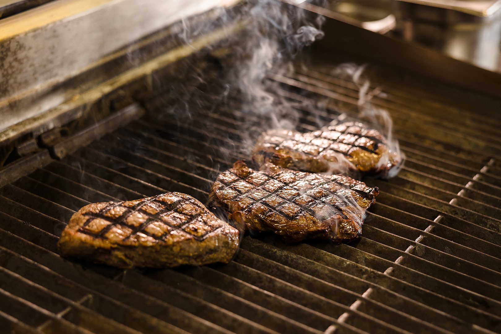 Three grilled steaks with sear marks cook on a commercial grill—perfect after a round of September golf, smoke rising.